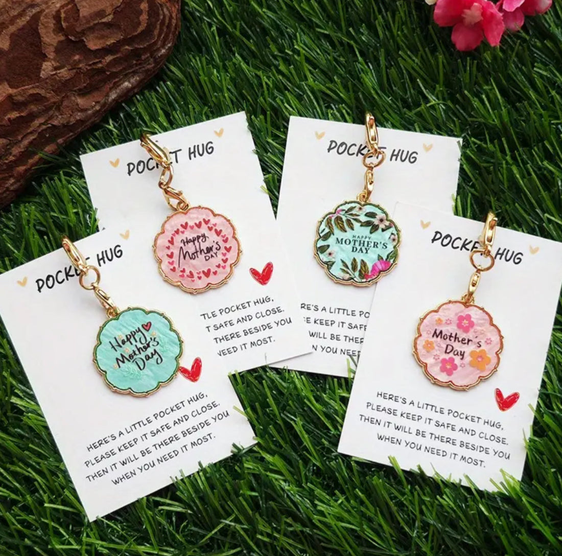 Three enamel keychains with messages 'Happy Mother's Day', 'Thinking of You', and 'Love You Mum', displayed on a grass-like surface, each with a colorful floral design on a round tag attached to the keychain.