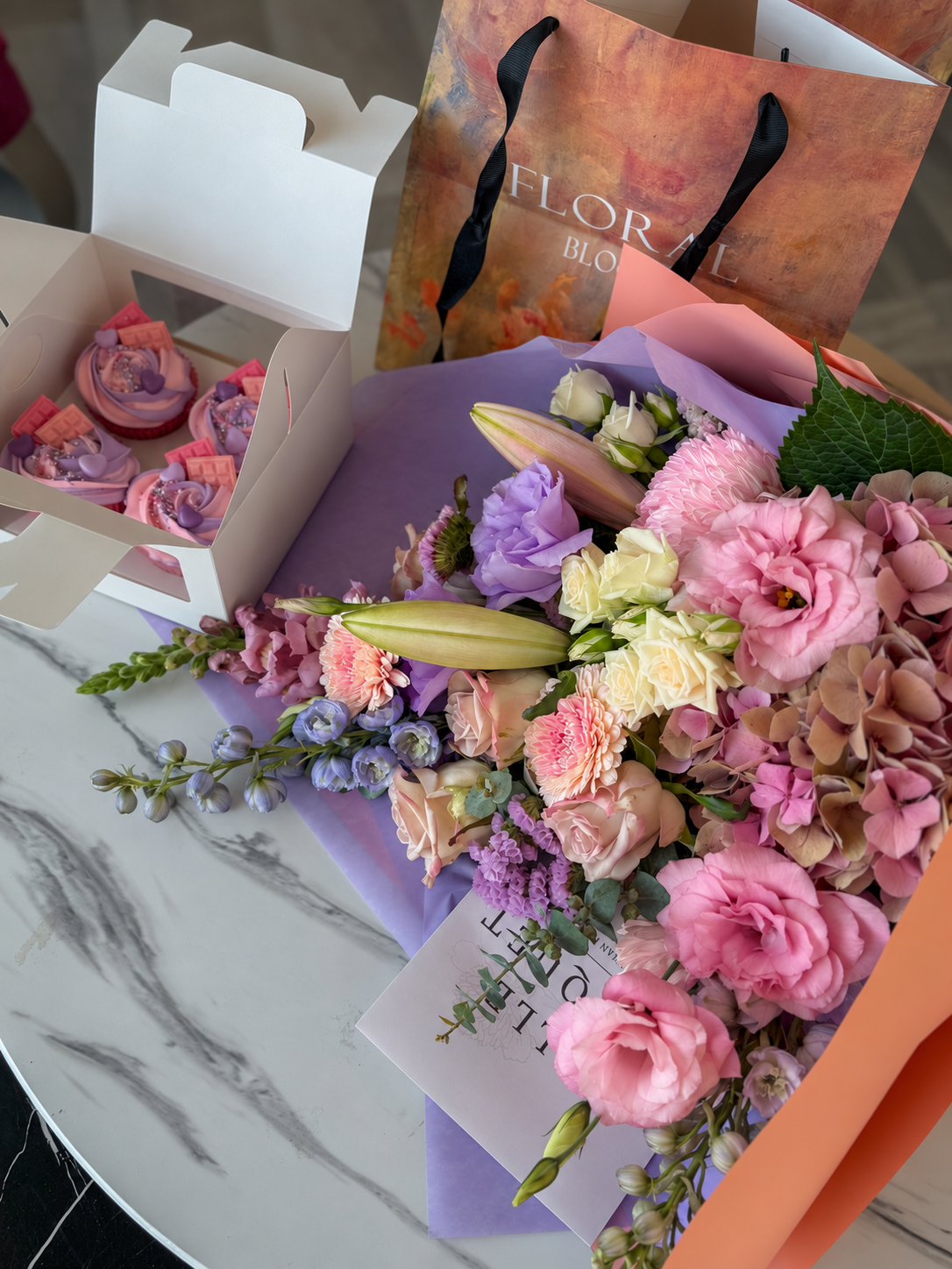 Angled view of a pink, lilac and cream bouquet with four decorated cupcakes, photographed in natural light