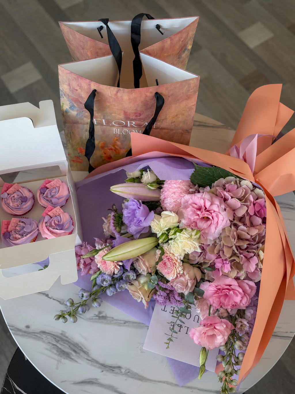 Pastel bouquet and four pink and lilac cupcakes on a marble table, with natural colours and soft shadows