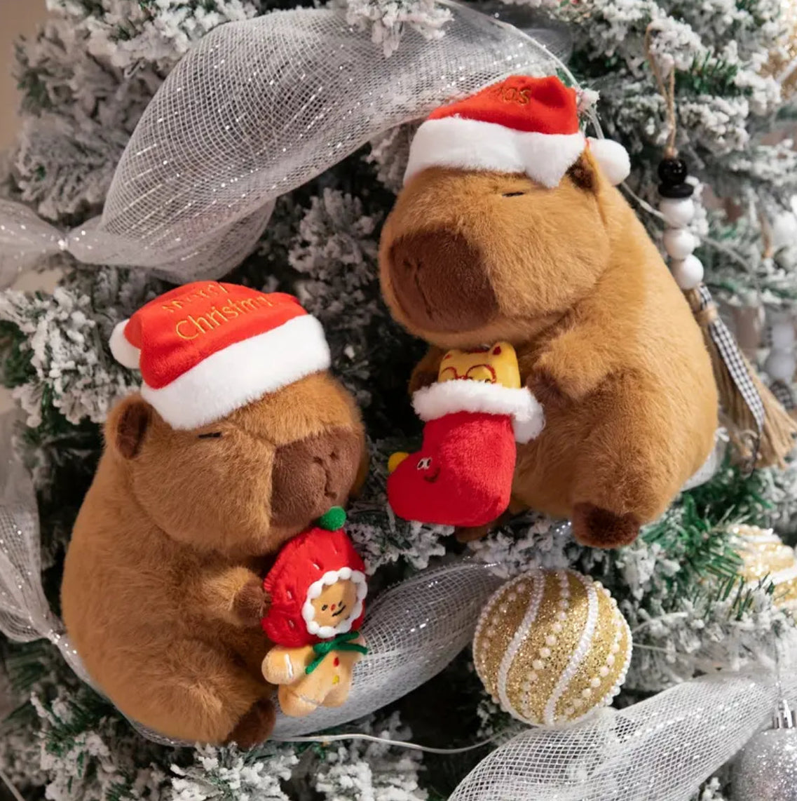 Two plush capybaras wearing Santa hats on a Christmas tree with ornaments.